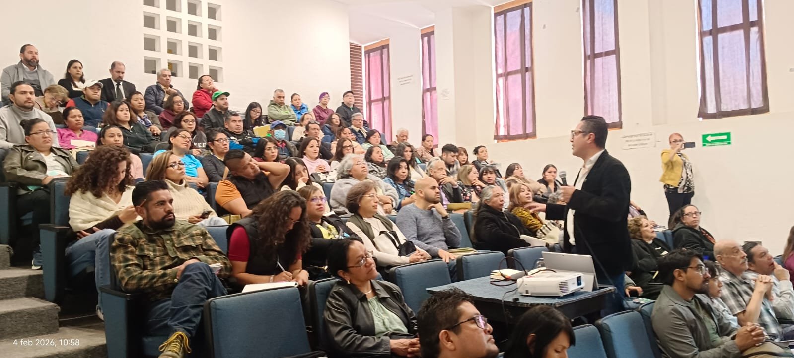 Large lecture hall filled with students seated in tiered rows listening to an instructor standing at the front of the classroom